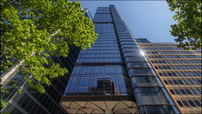 Looking up at glass wall skyscraper surrounded by green trees.